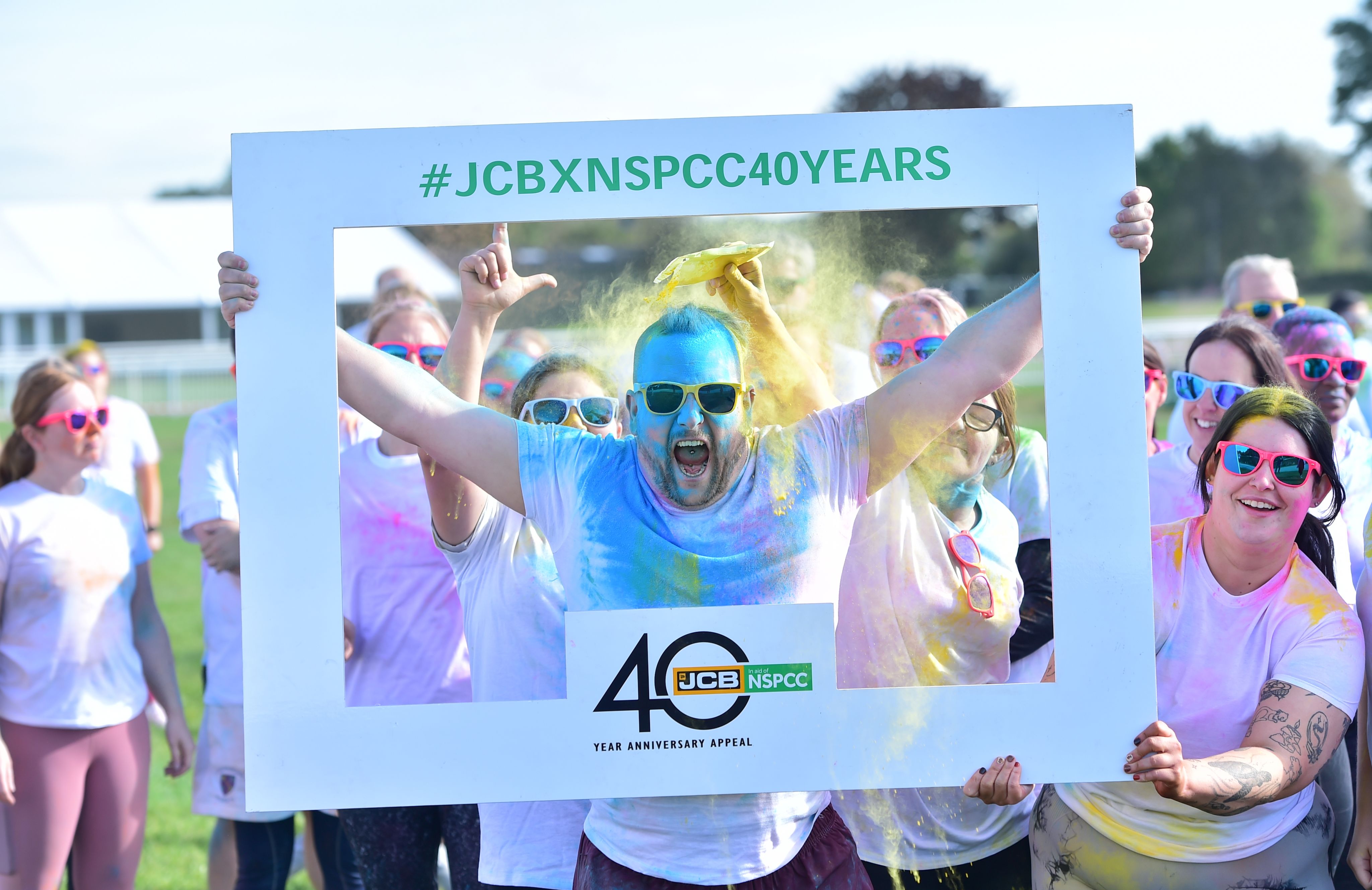 A group of people wearing brightly coloured sunglasses, covered in bright colours, holding a selfie frame that says 'JCBXNSPCC40YEARS'